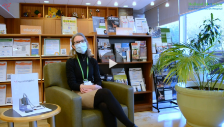 Sheri, an Occupational Therapist, is sat in front of a set of books on a green couch next to a large green plant.